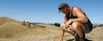 A man inspecting dry brown grass, with rolling brown hills and blue sky in the background.