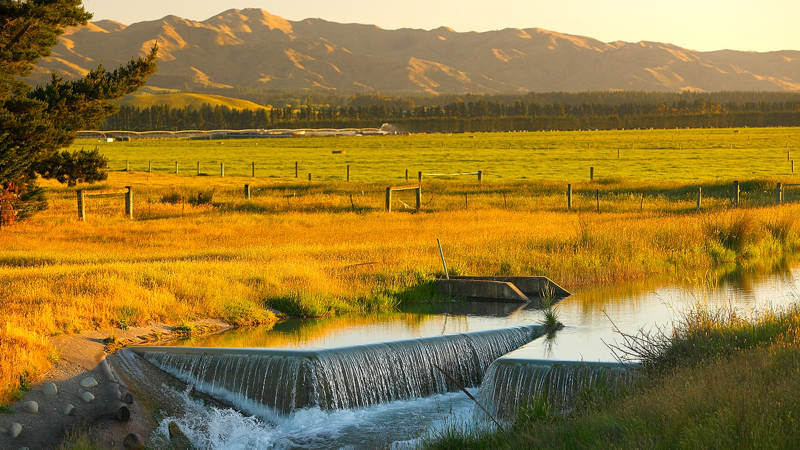 A rural scene with flat farmland and dry hills behind. In the foreground, water cascades over a small weir.