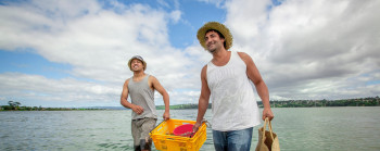 Two men smiling, one carrying a woven bag and both carrying a basket together as they wade through a body of water.