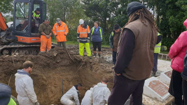 People beside and at the bottom of a large hole with a digger behind. Gravestones are in the foreground.