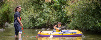 An older kid standing in a river, helping a young kid who is sitting in an inflated rate with a paddle.