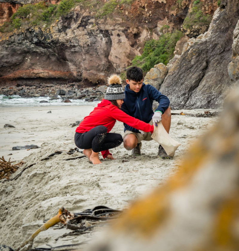 A boy and a girl crouching on a beach collecting rubbish. 