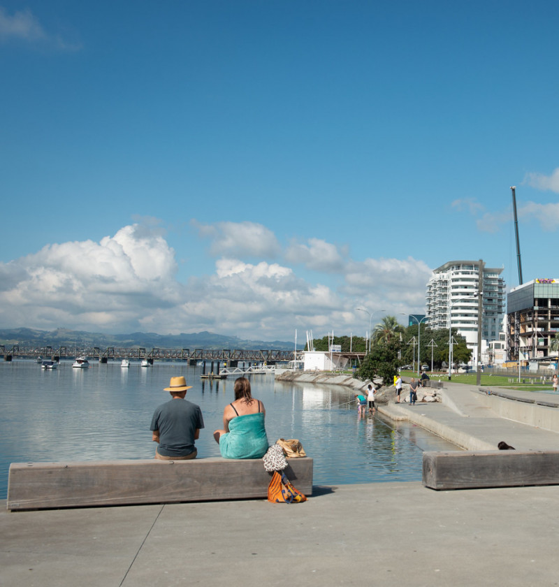 Two adults sitting by a harbour showing the ocean and city buildings. 