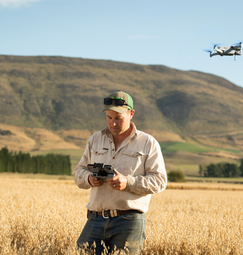 A farmer standing in a field holding a drone control. Farm hills can be seen in the background. 