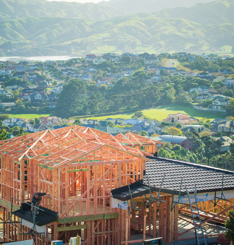 The framing of a new two-story house, with other suburban houses in the background. 