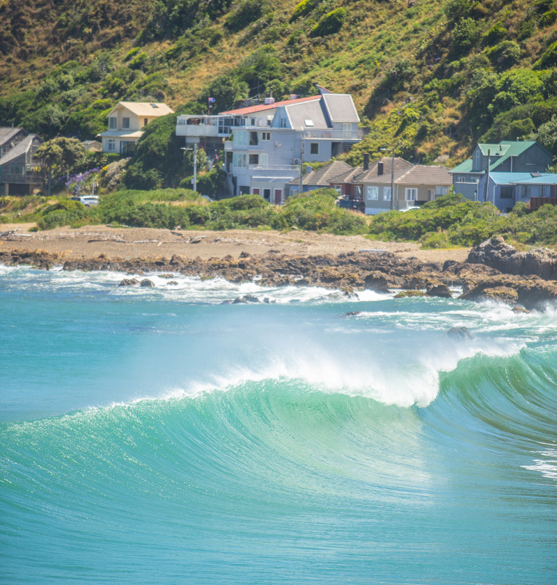 Houses by the beach, with green hills in the background and waves crashing on the rocks in the foreground.