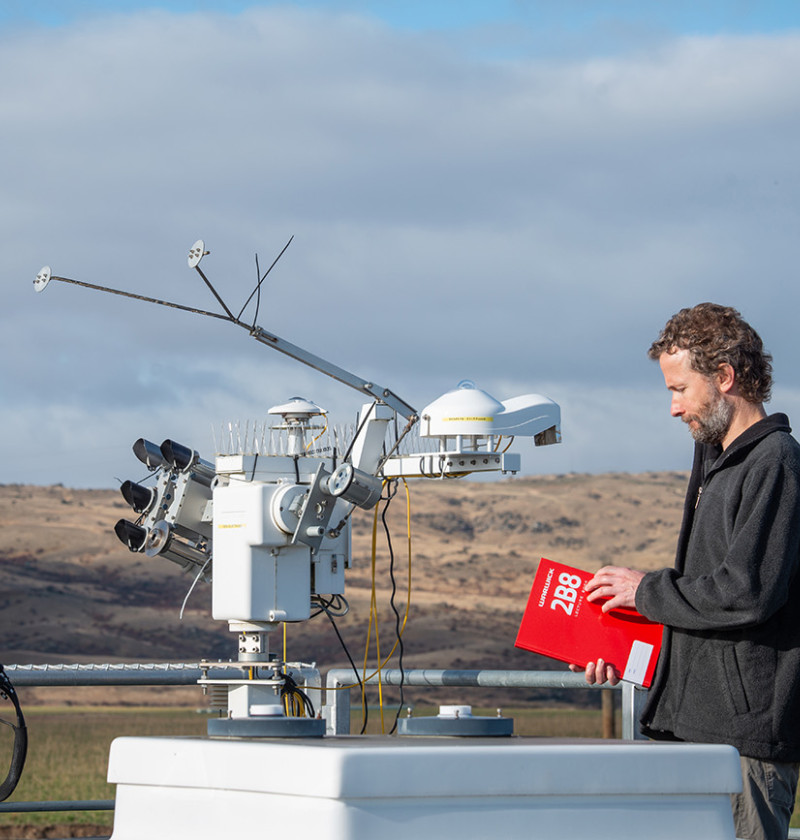 A man holding a notebook is standing by a piece of technology in a large field.