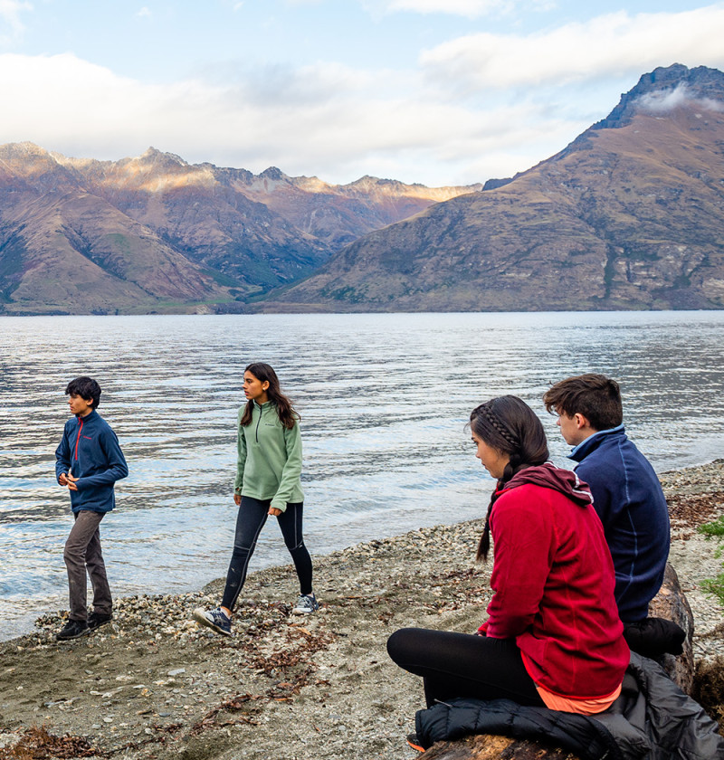 Two young adults are walking on the shore by some pristine water and two other young adults are sitting on a log on the shore. Mountains are in the background.