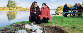 An adult and child smile whilst watching over a hāngī by the side of a river.