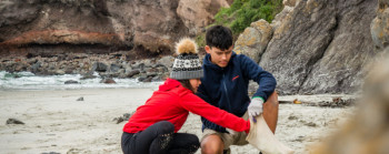 A boy and a girl crouching on a beach collecting rubbish. 