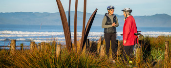 Two women talking next to a sculpture near the seaside. 