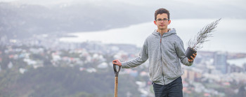 A teenage boy leaning on a shovel and holding a native tree. A city can be seen behind him.  