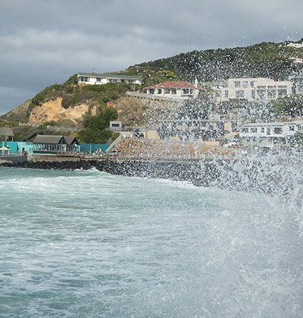 Two people walking alongside a waterfront, with big waves splashing up on them. Houses on hills are in the background.