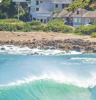 Houses by the beach, with green hills in the background and waves crashing on the rocks in the foreground.