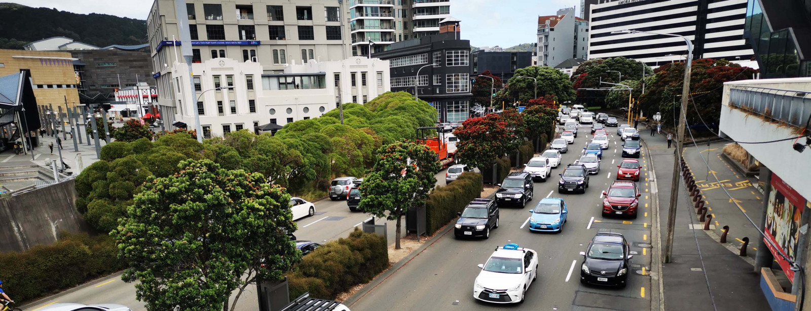 Photo of many cars stuck in slow moving traffic, in central Wellington. There are many tall buildings surrounding the busy six lane road, and some trees adjacent to the road. 