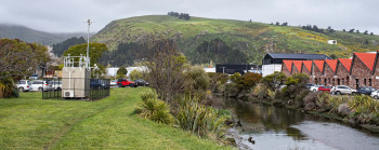 Photo of an air quality monitoring station, a small building with many sensors and aerials on the roof. It is located on a patch of grass next to a river, with industrial buildings and hills in the background.