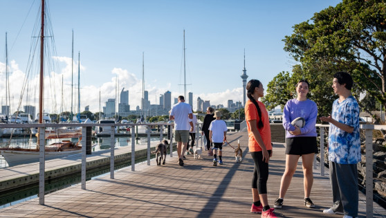 In the foreground, three young adults stand on a walkway, chatting. In the mid-ground, a family are walking their dogs. In the background are trees, Auckland's sky tower, buildings and the viaduct.